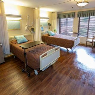 A clean and well-lit hospital room featuring two patient beds with large windows providing natural light.