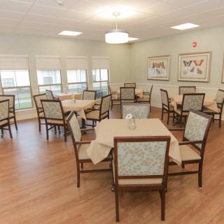A neatly arranged dining room with multiple tables and chairs, featuring soft lighting and large windows.