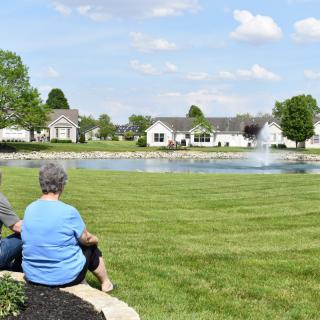 Two people sitting near a pond with a fountain, surrounded by grass and houses.