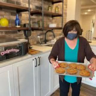 A person wearing a mask holds a tray of freshly baked cookies in a kitchen.