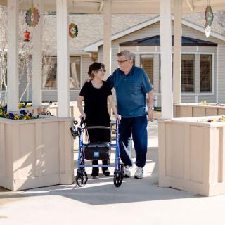 A caregiver assists an elderly person using a walker outside a building.