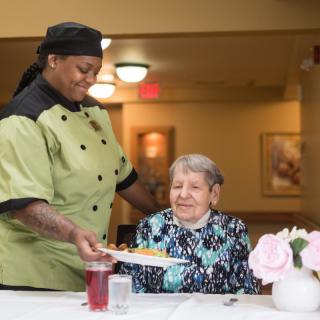 A chef serving a meal to an elderly woman in a dining setting.
