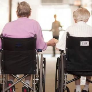 Two elderly individuals in wheelchairs holding hands in a care facility.