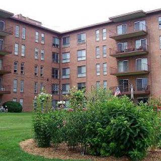A brick apartment building with balconies overlooking a well-maintained garden and lawn.