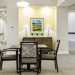 A well-lit room with a table and four chairs, featuring wall art and two lamps on a sideboard.