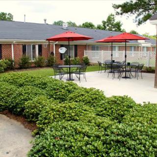 A cozy outdoor patio area with tables, chairs, and red umbrellas surrounded by shrubs.