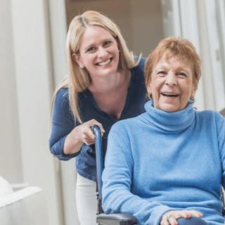 A caregiver happily assisting an elderly woman in a wheelchair indoors.