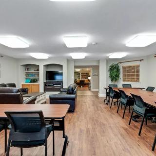 A spacious meeting room equipped with multiple tables and chairs, featuring a television and bookshelves against a backdrop of wooden flooring.