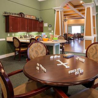 A cozy recreation room featuring a round table set up for a game of dominoes, with a kitchen area in the background.