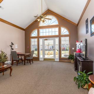 A cozy living room with large windows, a table and chairs, a television, and decorative plants.