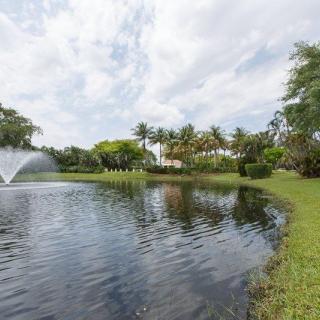 A picturesque view of a pond with a fountain surrounded by lush greenery and trees under a partly cloudy sky.