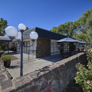A modern building with a stone wall and patio area, featuring lamp posts and surrounded by trees under a clear blue sky.