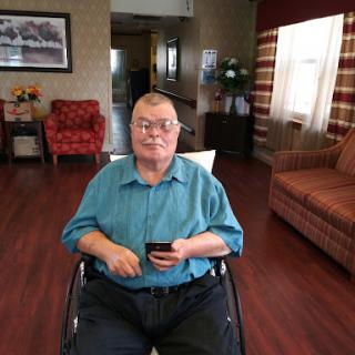 An elderly person seated in a wheelchair holding a smartphone, surrounded by a cozy living space.