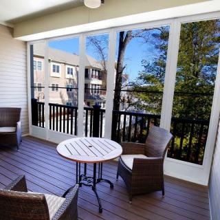 A cozy screened porch with outdoor furniture, featuring a round table and wicker chairs, overlooking trees and nearby buildings.