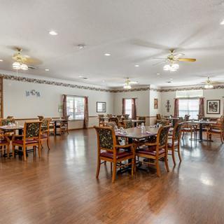 A spacious dining room with wooden floors and multiple tables and chairs, featuring ceiling fans for ventilation.