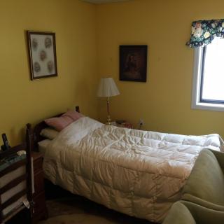 A cozy bedroom with yellow walls, featuring a single bed, artwork on the walls, a lamp, and a window with floral curtains.