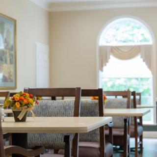 A cozy dining area with tables and chairs, featuring floral arrangements and a large window letting in natural light.