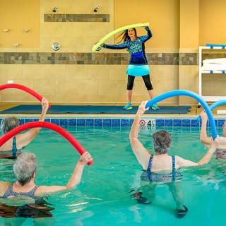 A group of seniors participating in a water aerobics class, using colorful noodles in an indoor swimming pool.