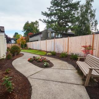 A landscaped garden with a winding concrete pathway and a wooden bench, bordered by a wooden fence.