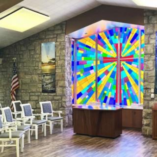 A chapel interior featuring a vibrant stained glass window with a cross, surrounded by chairs and stone walls.