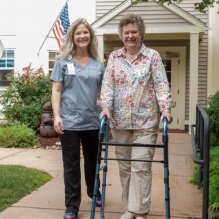A nurse assists an elderly woman using a walker outside a residential home.