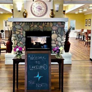 A welcoming lobby with a rustic stone fireplace, a clock above it, and decorative flowers on either side of a greeting sign.