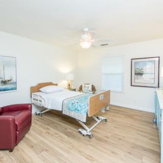 A clean and well-lit hospital room featuring a single adjustable bed, a red chair, framed artwork on the walls, and a wooden floor.