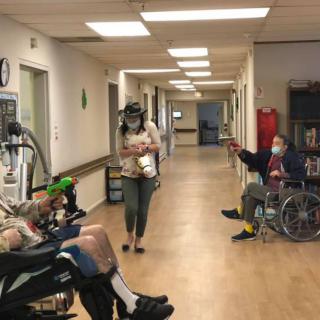 A hallway in a nursing home with residents in wheelchairs and a person walking by.