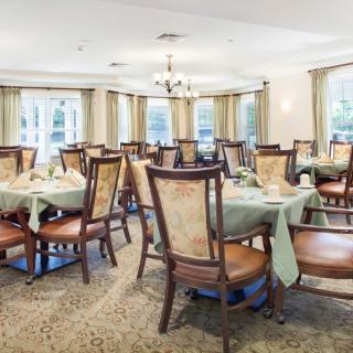 An elegant dining room with neatly arranged tables and chairs, featuring large windows and a central chandelier.
