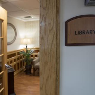 A view into a library through a wooden door, showing an interior with a lamp, plant, and seating area.