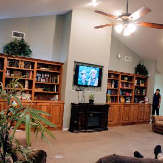 A cozy living room with wooden bookshelves, a mounted television, and a ceiling fan.