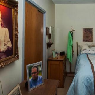 A cozy bedroom featuring a large framed portrait on the wall, a photo frame on a dresser, and a neatly made bed beside a coat rack.