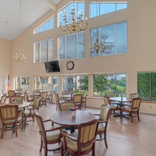 A spacious dining area with large windows, featuring multiple tables and chairs, chandeliers, and a wall-mounted television.