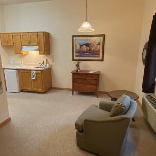 A cozy living space with a kitchenette, armchair, table, and wall painting.