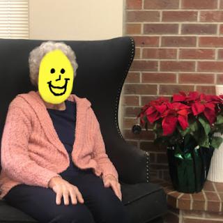 A cheerful elderly woman sitting comfortably in a chair, surrounded by festive potted plants.