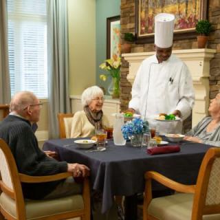 A chef serves a meal to elderly diners at a well-set table.