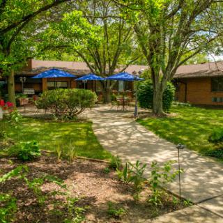 A peaceful garden setting featuring a winding pathway, lush greenery, and blue umbrellas providing shade.