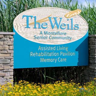 A sign for 'The Weils: A Montefiore Senior Community' offering assisted living, rehabilitation pavilion, and memory care services.