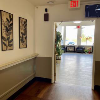 A hallway with artwork on the wall, leading to a bright area with seating and plants.