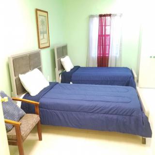 A tidy bedroom featuring two single beds with blue bedding, a window with curtains, a framed picture on the wall, and a chair by the bedside.