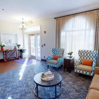 A cozy living room with patterned armchairs, a central coffee table, and sunlight streaming in through large windows.