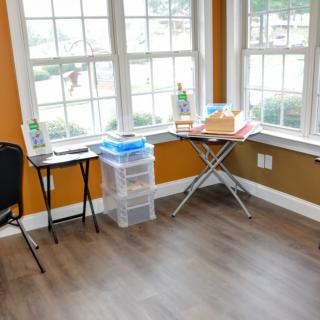 A bright room with large windows, featuring a small table, storage boxes, and a chair on a wooden floor.