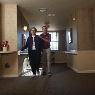 An elderly couple walking together in a hallway, with one providing support to the other.