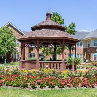 A wooden gazebo surrounded by vibrant flowers in a garden setting with buildings in the background.