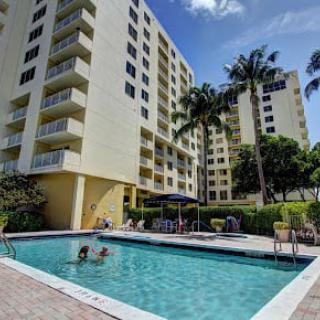 An outdoor swimming pool surrounded by apartment buildings and palm trees under a clear blue sky.