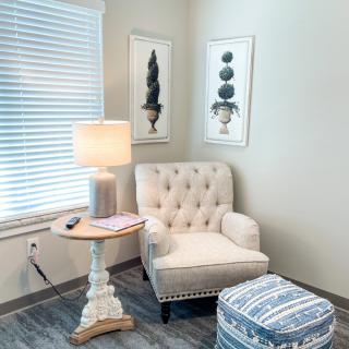 A cozy corner featuring a tufted armchair with a side table and lamp, complemented by artwork on the walls.