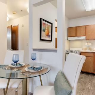 A modern apartment dining area with a glass table set for two, adjacent to a compact kitchen.