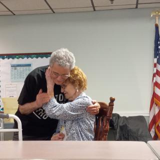 An elderly couple shares a heartfelt embrace in a room with an American flag in the background.