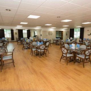 A spacious dining hall with wooden flooring, featuring multiple tables and chairs arranged neatly.
