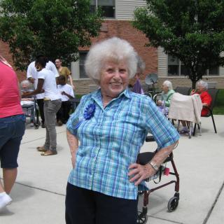 An elderly woman poses confidently at an outdoor gathering, with others socializing in the background.